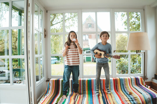 Siblings playing musical instrument standing on sofa at home