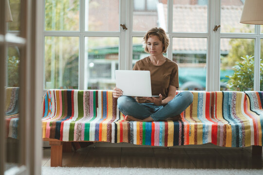 Woman using laptop sitting on sofa in front of window