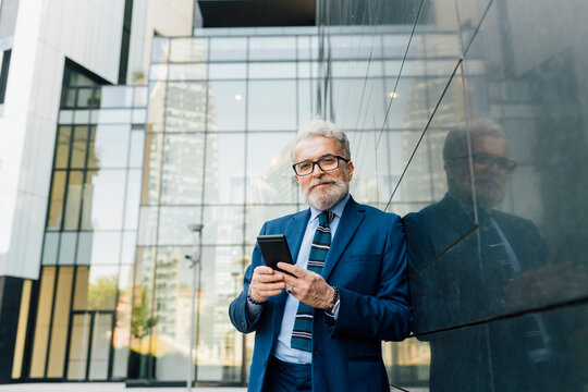 Smiling businessman standing with smart phone by wall outside office building