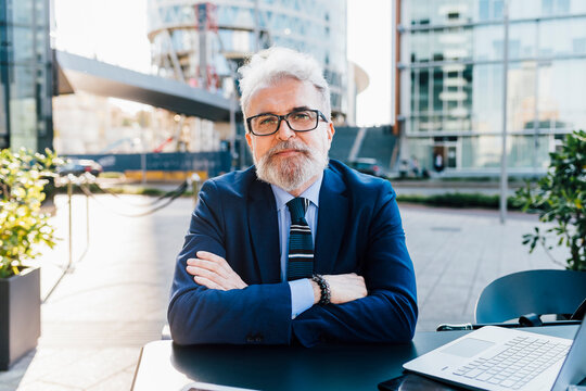 Senior businessman with arms crossed sitting by laptop outside building