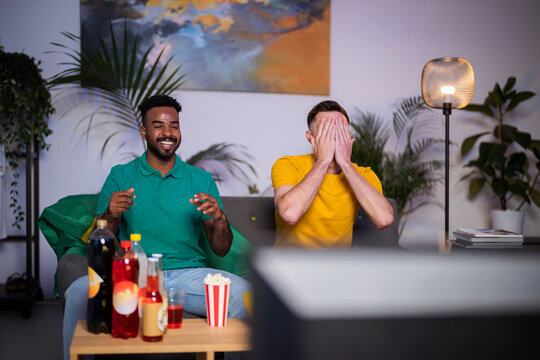 Sad man covering face sitting by happy friend watching football match at home