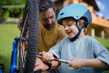 Father with little son together preparing bicycle for a ride, pumping up tyres in garden in front...