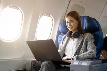 Asian young woman wearing headphone using laptop sitting near windows at first class on airplane...