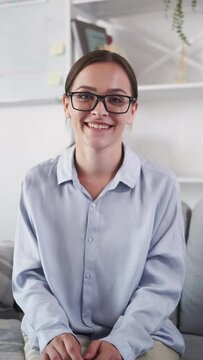 Online interview. Video call. Digital conference. Cheerful smiling smart woman in glasses listening nodding discussing on couch at light home interior vertical.