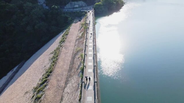 A dynamic high-angle aerial shot of the bridge of the Shing Mun Reservoir dam in Hong Kong. There are tourists walking along the pathway across the dam and the sunlight reflects on the water's surface