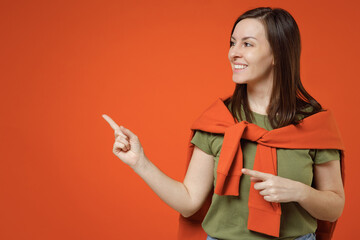 Young smiling happy woman 20s wearing khaki t-shirt tied sweater on shoulders point index finger aside on workspace area mock up isolated on plain orange background studio. People lifestyle concept.