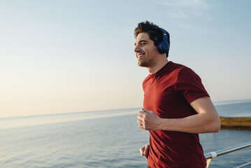 Side view young strong sporty athletic fit sportsman man wear sports clothes heapdhones listen music warm up training at sunrise sun dawn over sea beach outdoor on pier seaside in summer day morning.