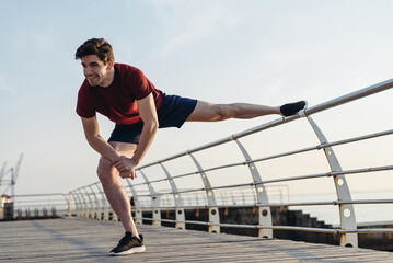 Full length side view young strong sporty fit sportsman man 20s in sports clothes do stretch exercise for legs warm up training at sunrise sun dawn over sea beach outdoor on pier seaside in morning