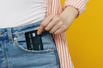 Cropped photo shot close up woman she wears striped shirt white t-shirt hold put into pocket mock up of credit bank card isolated on plain yellow background studio portrait. People lifestyle concept.