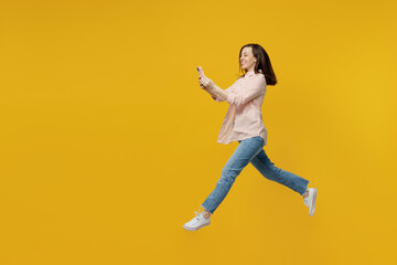 Full body side view young fun woman she 30s in striped shirt white t-shirt jump high run fast hurry up hold use mobile cell phone isolated on plain yellow background studio. People lifestyle concept