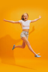 Fototapeta na wymiar Thrilled young girl with long blond hair wearing white t-shirt jumping isolated on yellow background. Concept of emotions, facial expression, youth, fashion