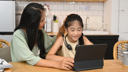 Obraz premium Adorable little asian girl doing homework, learning online at home and her mother sitting nearby and giving support