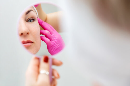 Cosmetology and plastic surgery. Close up of reflection of a woman looking at herself in the mirror. The beautician tightens the client's eyebrow