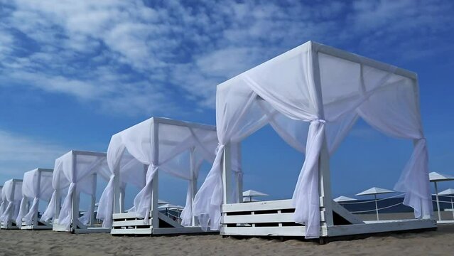 Wooden beach canopies on the blue sky background. The white curtains of the awnings flutter in the sea breeze.