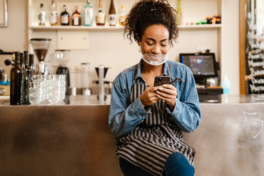 Young black waitress in face mask using mobile phone at cafe