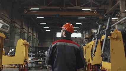 Rear tracking of workman in orange hat walking at heavy machinery production plant
