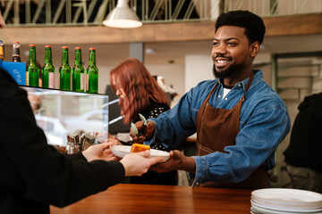 Black bearded man wearing apron smiling while working in cafe