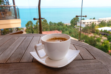 White cup of coffee on a wooden table on the balcony with a blurred background of the sea, green trees, sky