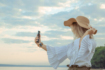 Portrait of a girl taking a selfie on the smartphone. Young beautiful 30 years woman caucasian appearance in straw hat with satin ribbon bow. Copy space. Summer lifestyle concept. Natural background