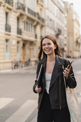 cheerful woman in black leather jacket holding laptop and smartphone on street in paris.