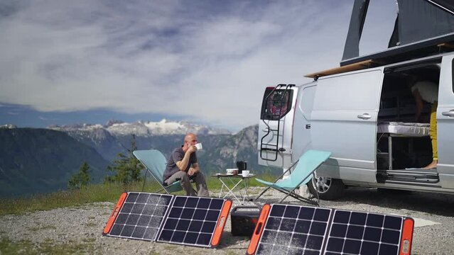 Mature adult sporty couple sitting in camping chairs high up in the mountains close to their camper van having breakfast on sunny morning, self sufficient with portable solar panels, woman getting out