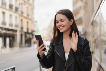 pleased young woman in stylish jacket waving hand while having video chat on street in paris.