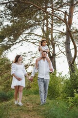 Fototapeta premium Beautiful pregnant brunette mom and dad and dauther in white clothes look at each other in nature.