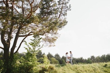 Beautiful pregnant brunette mom and dad and dauther in white clothes look at each other in nature.