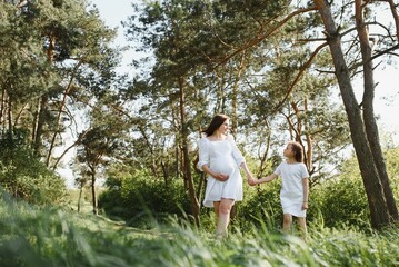 Happy family: a young beautiful pregnant woman with her little cute daughter on a sunny summer day. Parents and kids relationship. Nature in the country.