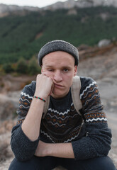 Boy with hat and backpack sitting on a rock with a tired face in the middle of mountains