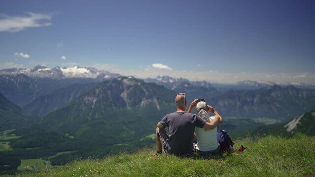 Mature adult sporty couple sitting together in alpine meadow high up in european alps enjoying view at austrian mountains with dachstein glacier in background on sunny summer day taking selfie picture
