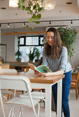 Young student brunette woman reading a book and studying at cafe with modern green interior design. Female student at coworking space or library or cafe, back to school education concept