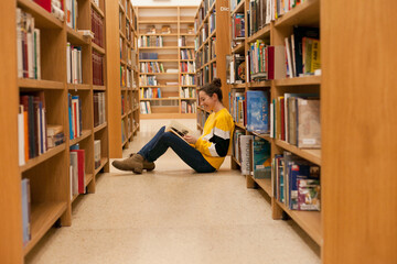 Young female student sitting on the floor near bookshelf reading a book. Woman wearing jeans and yellow sweatshirt studying while sitting in college library or bookstore. 