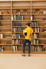 Young female student reading a book from wooden bookshelves in university library. Woman in public library inside the building searching for books.. 
