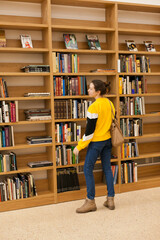 Young female student taking a book from wooden bookshelves in university library. Woman in public library inside the building searching for books.. Woman in jeans and yellow sweatshirt at bookstore.