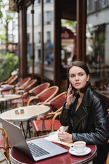 young freelancer in black jacket talking on smartphone near laptop and cup of coffee on table in french outdoor cafe.