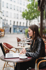 side view of smiling freelancer in black jacket looking at laptop near cup of coffee on table in french outdoor cafe.
