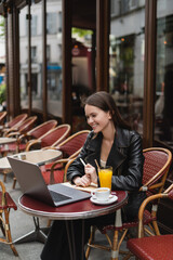 happy freelancer in black leather jacket looking at laptop near drinks on table in french outdoor cafe.
