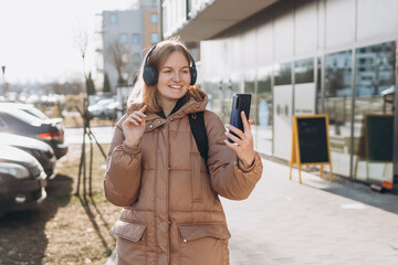 Portrait of young happy woman walking on the street in the city. Girl talking on a video call with phone and headphones outdoor. Young woman listening music in headphones in the city.