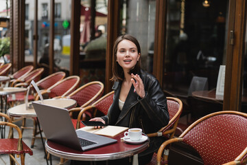 young freelancer in black jacket holding smartphone and waving hand near laptop and cup of coffee in french outdoor cafe.