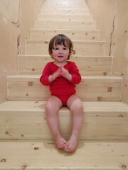 Funny Toddler Baby girl in red body at sits on stairs indoors