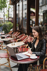happy freelancer in black jacket having video call near cup of coffee on table in french outdoor cafe.