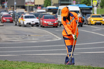 Woman worker in orange uniform with a broom sweeps the road on cars background. Street cleaning in...