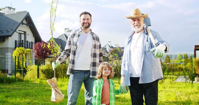 Portrait shot of cute small boy with happy father and grandfather smiling to camera in garden. Male genarations of family planting trees at summerhouse. Outside.