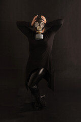 A model girl in a gas mask, leather or latex pants, boots and robes stands on a black background in a pretentious pose. Fetish, BDSM, perversion or gas attack, bio o radiation hazard - obrazy, fototapety, plakaty