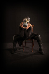 A girl in a full gas mask, latex pants, boots and robes is sitting on a chair on a black background in a pretentious pose with her neck in her hands. Fetish, BDSM, perversion, bio o radiation hazard
