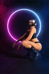 Seductive girl fetish model with a slim figure poses sitting in a black gas mask, black leather or latex sexy underwear and leather boots with neon lights on the background. bdsm, fetish, polluted air - obrazy, fototapety, plakaty