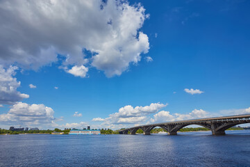 Fototapeta premium View from the right bank of the Dnieper river over the first metro bridge in Kyiv city with blue subway train, motorboat under it and hotelship behind.