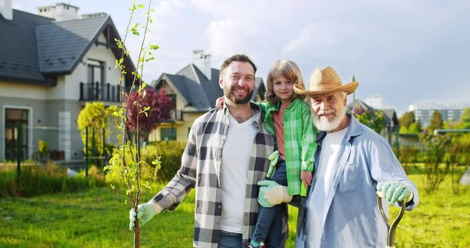 Portrait of happy three male genarations smiling to camera in garden on summer sunny day. Kid planting tree with father and grandfather. Senior man with adult son and little grandson.