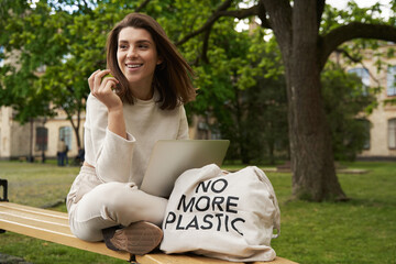 Cheerful woman sitting near eco bag and using notebook outdoors